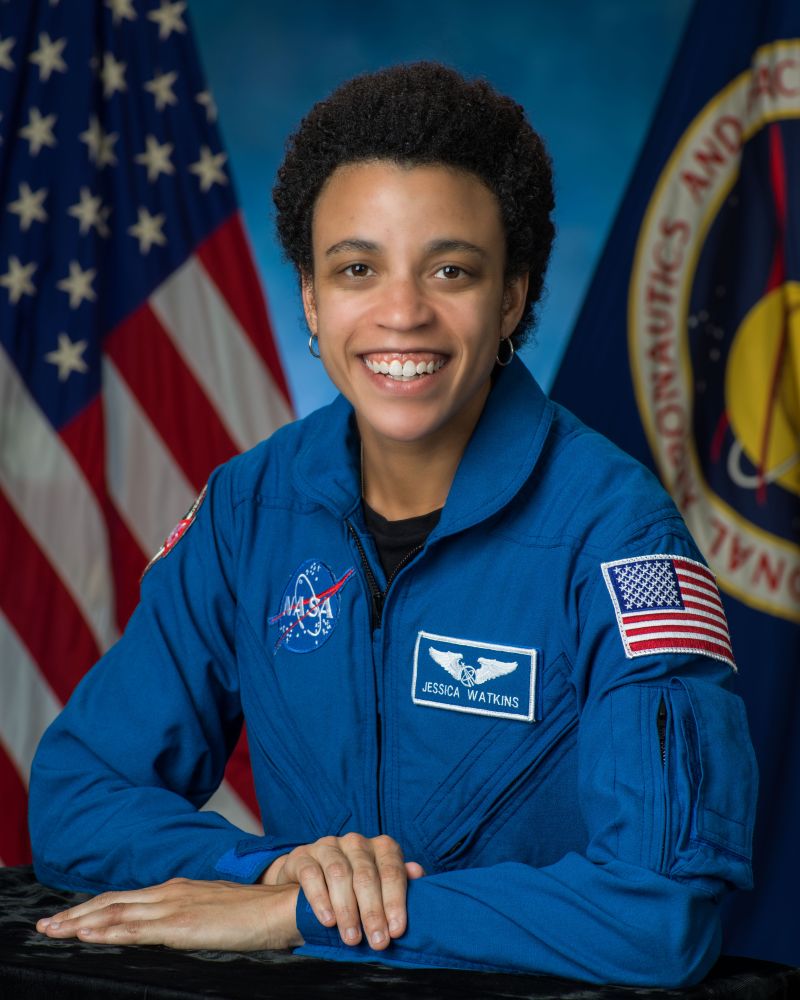 Jessica Watkins is smiling while wearing a blue NASA flight suit in front of an American flag