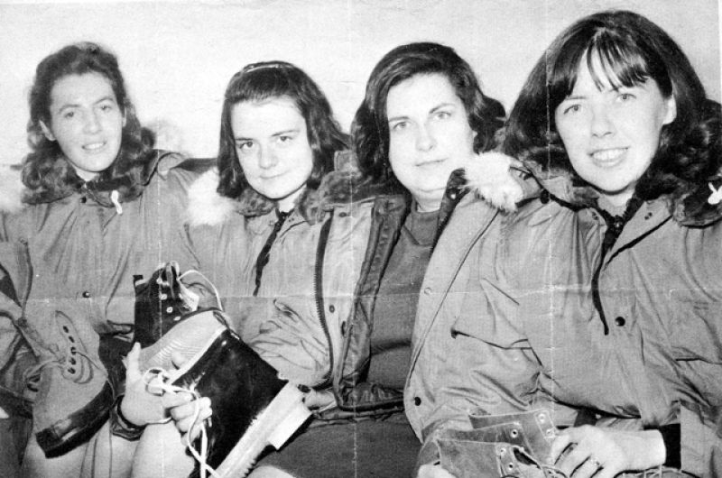 A black and white photo depicts four women in heavy coats sitting against a wall