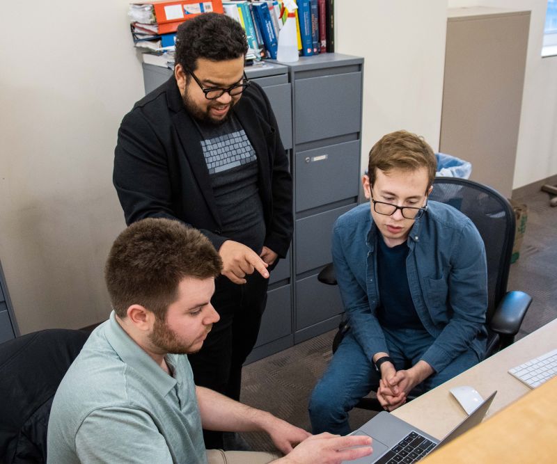 Three men looking at a laptop. One man is saying something to the others.
