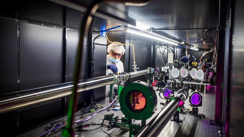 A man wearing protective gear adjusts a long metal tube inside a laser experiment chamber.