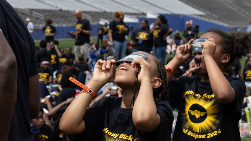 Two kids in matching eclipse t-shirts look up at the sky through paper eclipse glasses.