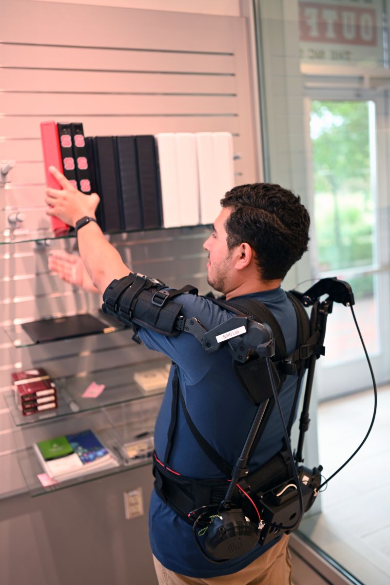 A man demonstrates an AI-powered soft-shoulder exosuit with improved manipulation capabilities. The suit supports his back and upper arms. He is reaching to pick something up from a shelf above his head.