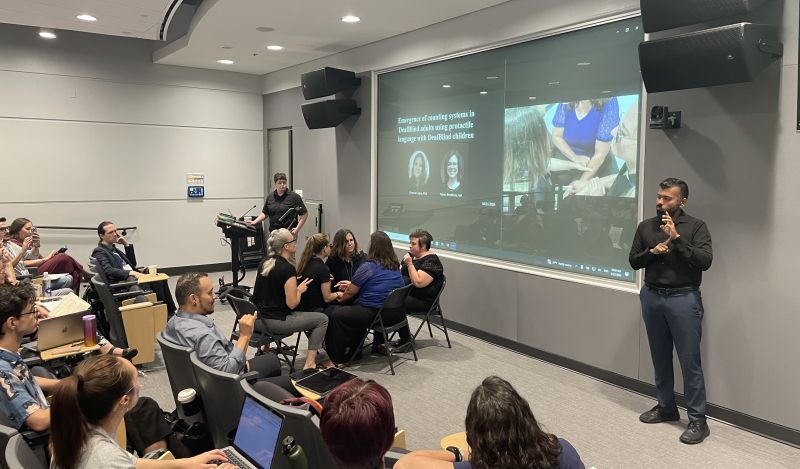 A DeafBlind presenter and her co-investigator are presenting in front of an audience at the MCLS conference. They sit at the front of the room with Protactile American Sign Language (PTASL) interpreters. An American Sign Language (ASL) interpreter is also seen standing at the front. The title of the presentation and a closeup video of how communication occurs in PTASL are projected on the screen behind the presenters.