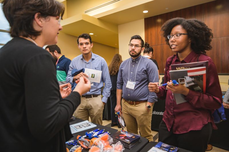 A person in the foreground stands at a table and speaks to several students who are holding pamphlets.