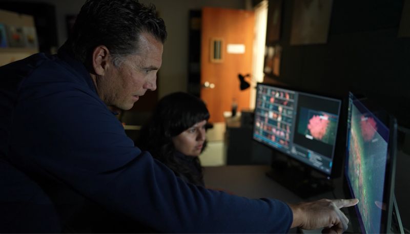 Two researchers in a dark room pointing at computer monitors.