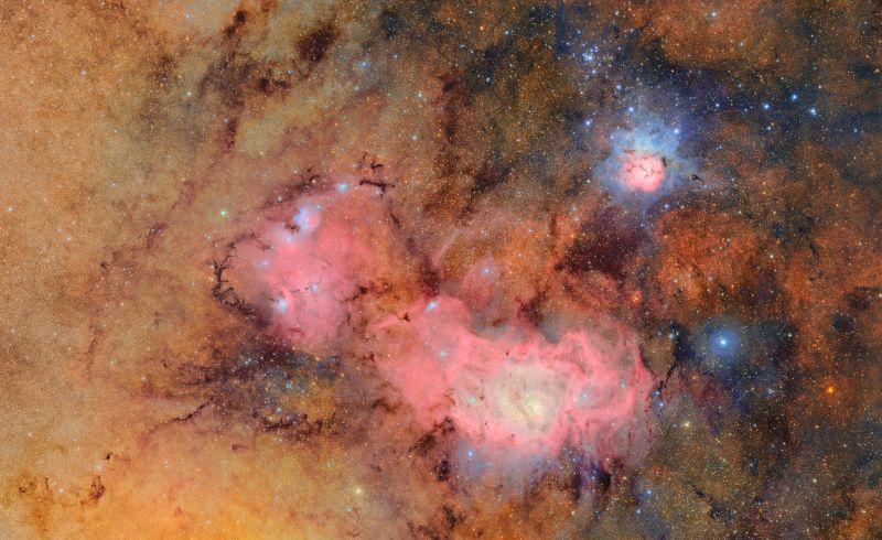 Against a background dense with glowing orange and golden stars, two nebulas appear as pinkish clouds of gas and dust.
