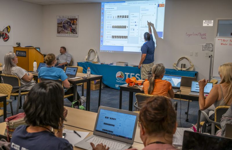 An instructor points to something projected on a screen while teachers look on from tables with laptop computers on them.