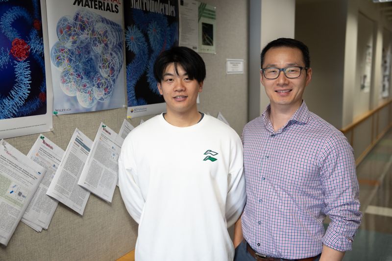 Two men stand in a hallway next to a billboard with scientific papers hanging on the wall.