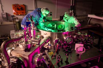 Two men in lab coats, hair nets, gloves and protective eyeglasses adjust the optics inside a circular metal tank. Metal tubes enter the tank from the sides, and more optical components are secured to a table outside.