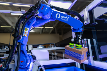 A blue robot arm, labeled "ambi robotics," holding a cardboard package.