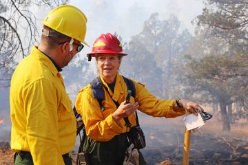 Mary Lata, a woman in a red hard hat and yellow hi-vis jacket speaks to a man in a yellow hard hat and yellow shirt. IN the background, a fire is smoldering.