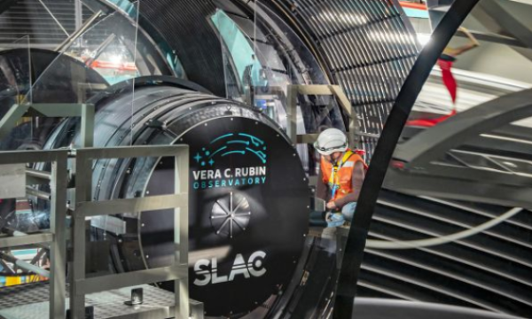 A worker, wearing a hard hat, kneels near a human-sized camera installed in an observatory.