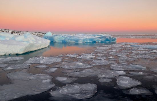 Ice floats on top of a body of water in front of a pink sky 