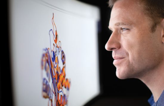 A man smiles slightly as he stares at a large computer screen that displays a multicolored model of a virus