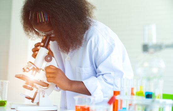 A young girl in a lab coat looks through a microscope