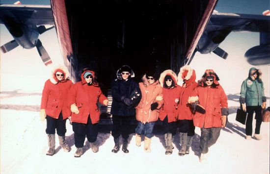 Six women in cold weather gear stand in the snow with a large aircraft behind them