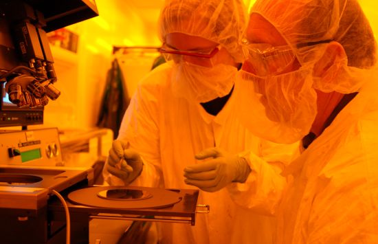 Researchers wearing lab protective gear work at a microscope in a room with an orange glow