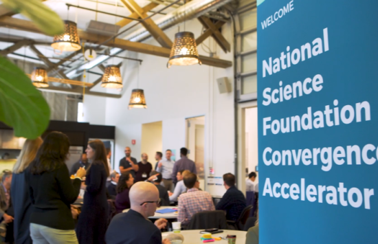 A group of people at a workshop with a sign that says "Welcome: National Science Foundation Convergence Accelerator"
