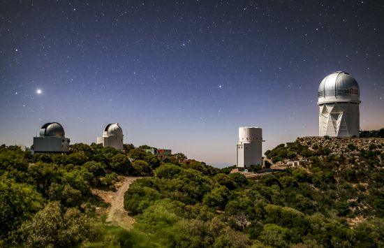 Nighttime scene of a hilly area with five buildings, including observatories, against a background of a star-filled sky.