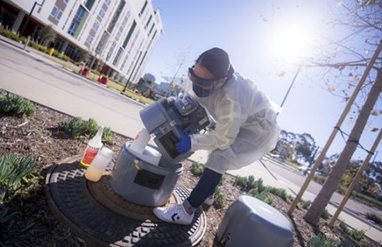 A researcher who is wearing protective labwear handles a wastewater sampler outdoors.