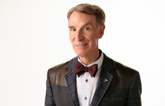 A man wearing a coat and bowtie against a white background smiles at the camera.