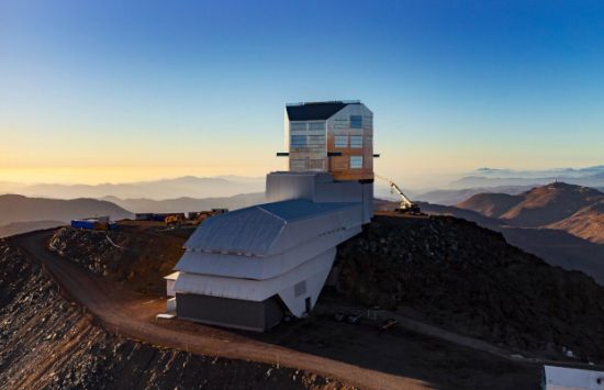 Rubin Observatory stands prominently in the center of this image atop its Chilean desert summit on Cerro Pachón. The sky is clear blue, and the setting sun glows from the left, illuminating the left side of the observatory. A large crane sits to the right of the observatory with its arm extended, looking small compared to the observatory building. The foreground summit area is shadowed, and the desert mountains recede into the distance, interspersed with hazy atmosphere.