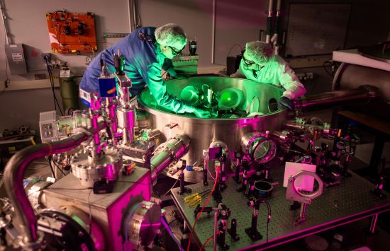 Two men in lab coats, hair nets, gloves and protective eyeglasses adjust the optics inside a circular metal tank. Metal tubes enter the tank from the sides, and more optical components are secured to a table outside.