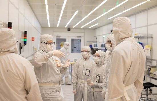 A group of researchers in white protective gear and masks stand in a brightly lit white-walled hallway.