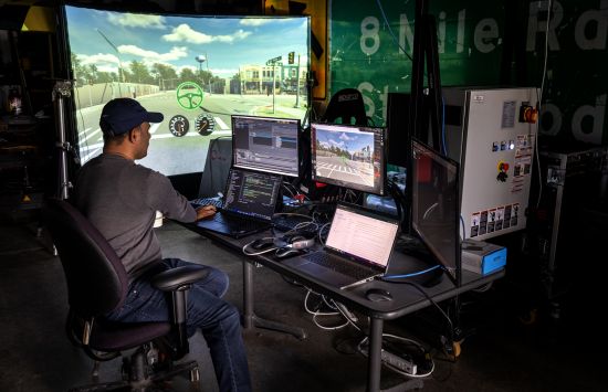 A person sitting at a computer in a darkened room in front of a screen where a simulated road is projected.