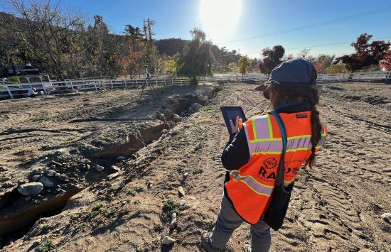 A researcher in a safety vest holding a tablet stands on the edge of a ditch.