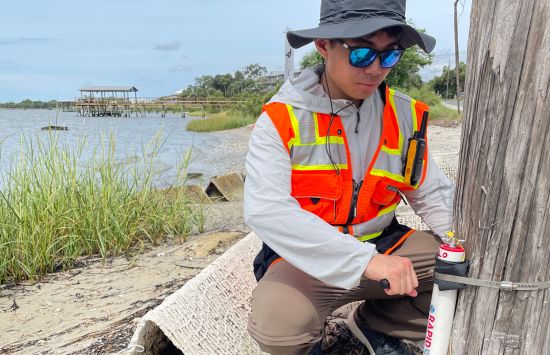A person in an orange visibility vest secures a data tracker to a utility pole near a shoreline.