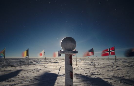 A sphere on a pedestal stands in the snow marking the Ceremonial South Pole with flags in a row waving in the background.