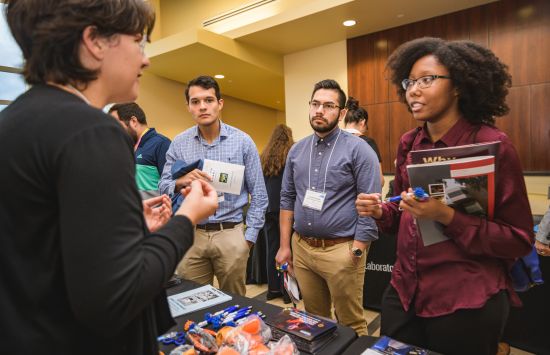 A person in the foreground stands at a table and speaks to several students who are holding pamphlets.