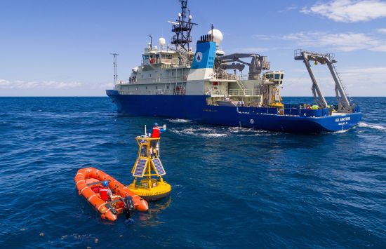An orange inflatable boat next to a yellow weather buoy with a large research vessel in the background.