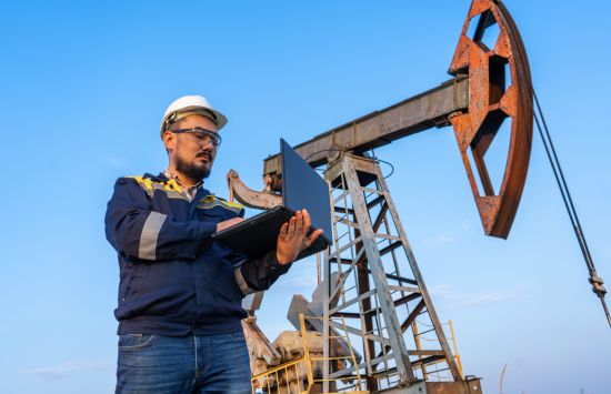 An engineer with a laptop in front of an oil rig.
