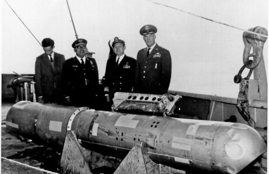 A black-and-white photograph of four men, three wearing military uniforms, standing behind a hydrogen bomb.