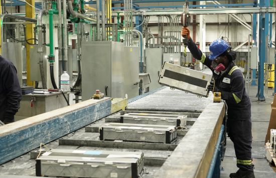 A factory worker, wearing a protective mask and helmet, suspends a large battery from a pulley.