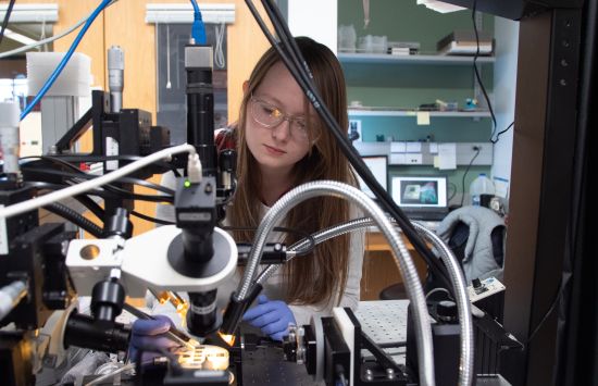 A student surrounded by equipment working on a specimen under a microscope.