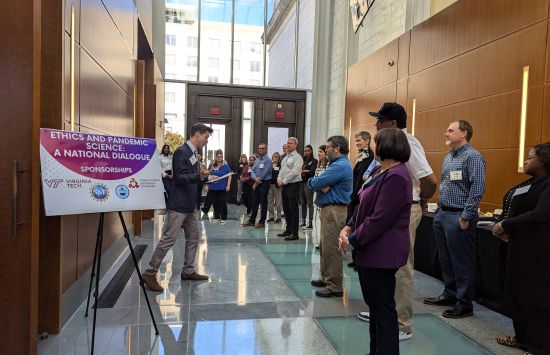 Several people stand in the hallway of a conference center. They appear to be waiting to enter a session titled "Ethics and Pandemic Science: A National Dialogue" as there is a sign outside the meeting room.