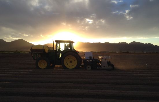 As the sun sets behind mountains, a tractor pulls a wheeled robot across dirt-covered fields.