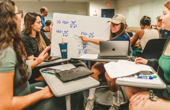 Students sitting at desks pulled together closely look at math equations on a white board.