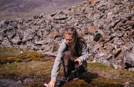 A person taking water samples from a stream with a mountain in the background.
