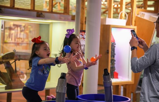 Two young girls play at an interactive museum exhibit, where plastic balls hover in the air above air-blowing tubes.