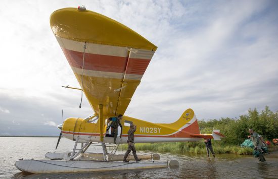 People loading items onto a yellow and orange float plane.