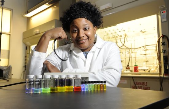 A researcher in a lab coat stands behind a collection of colorful vials of liquid.