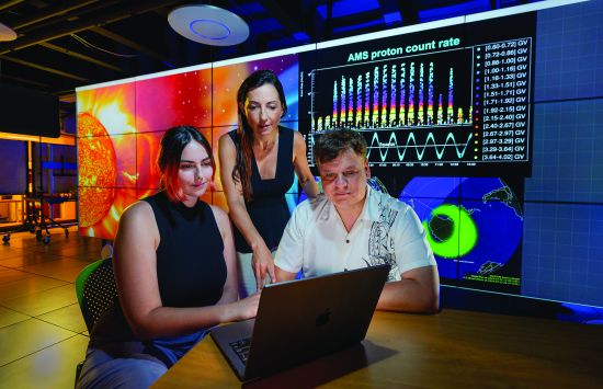Three people looking at a laptop in front of a large screen displaying a colorful graph.