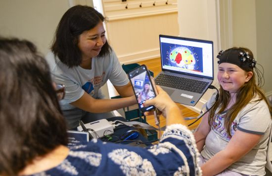 Lisa Odom of Alexandria, Va., takes a photo of her daughter, 10-year-old Josie, as she wears a functional Near-Infrared Spectroscopy (fNIRS) cap, while UMD human development doctoral student Gavkhar Abdurokhmonova holds a laptop showing the most active parts of her brain.