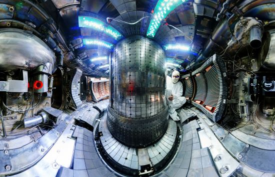 A photograph taken with a fisheye lens, featuring a researcher wearing protective gear and sitting within a chamber of metal, lights and gadgetry.