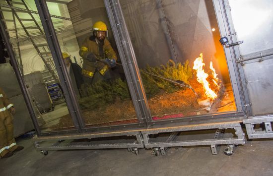 A firefighter stands in a glass walled wind tunnel, where he uses a hose to extinguish a fire ignited in grass.