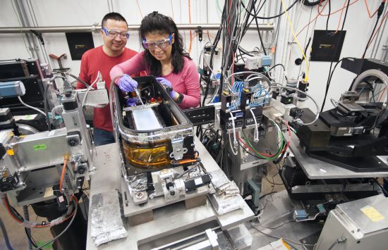 Two researchers in protective eyewear adjust an apparatus in a lab full of metal equipment and wiring.